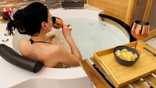 Eine Person entspannt in einer Badewanne, neben ihr ein Tablett mit Snacks und Getränken.
