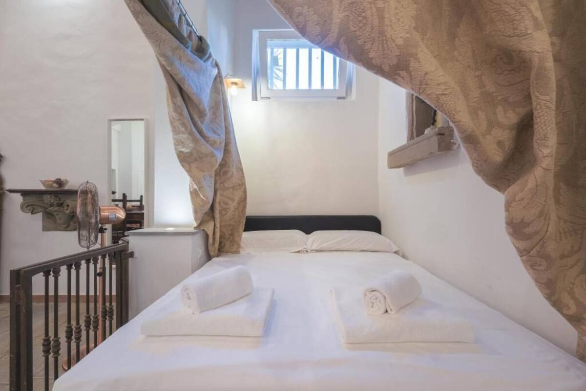 A hotel room with a bed, two rolled towels, a window, a mirror, and a small shelf with decorative items.