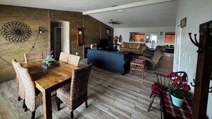 Un amplio salón con una mesa de comedor de madera, sillas de mimbre, un sofá y un sofá azul. Se ve una cocina al fondo.