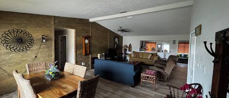 A spacious living area with a wooden dining table, wicker chairs, a sofa, and a blue couch. A kitchen is visible in the background.