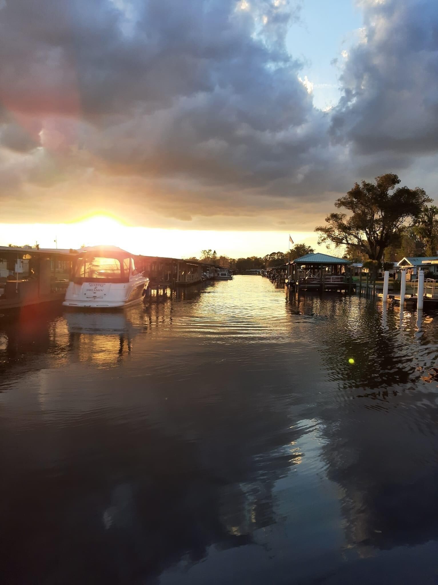Een rustige rivier met boten aan de linkerzijde, een steiger aan de rechterzijde en een zonsondergang op de achtergrond.