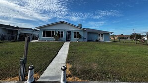 Una casa residencial con jardín delantero, un camino que lleva a la entrada y un cielo azul despejado con nubes.