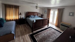 Un dormitorio con una cama de madera, una cama individual, un ventilador de techo, una ventana con cortinas y una puerta con vistas.