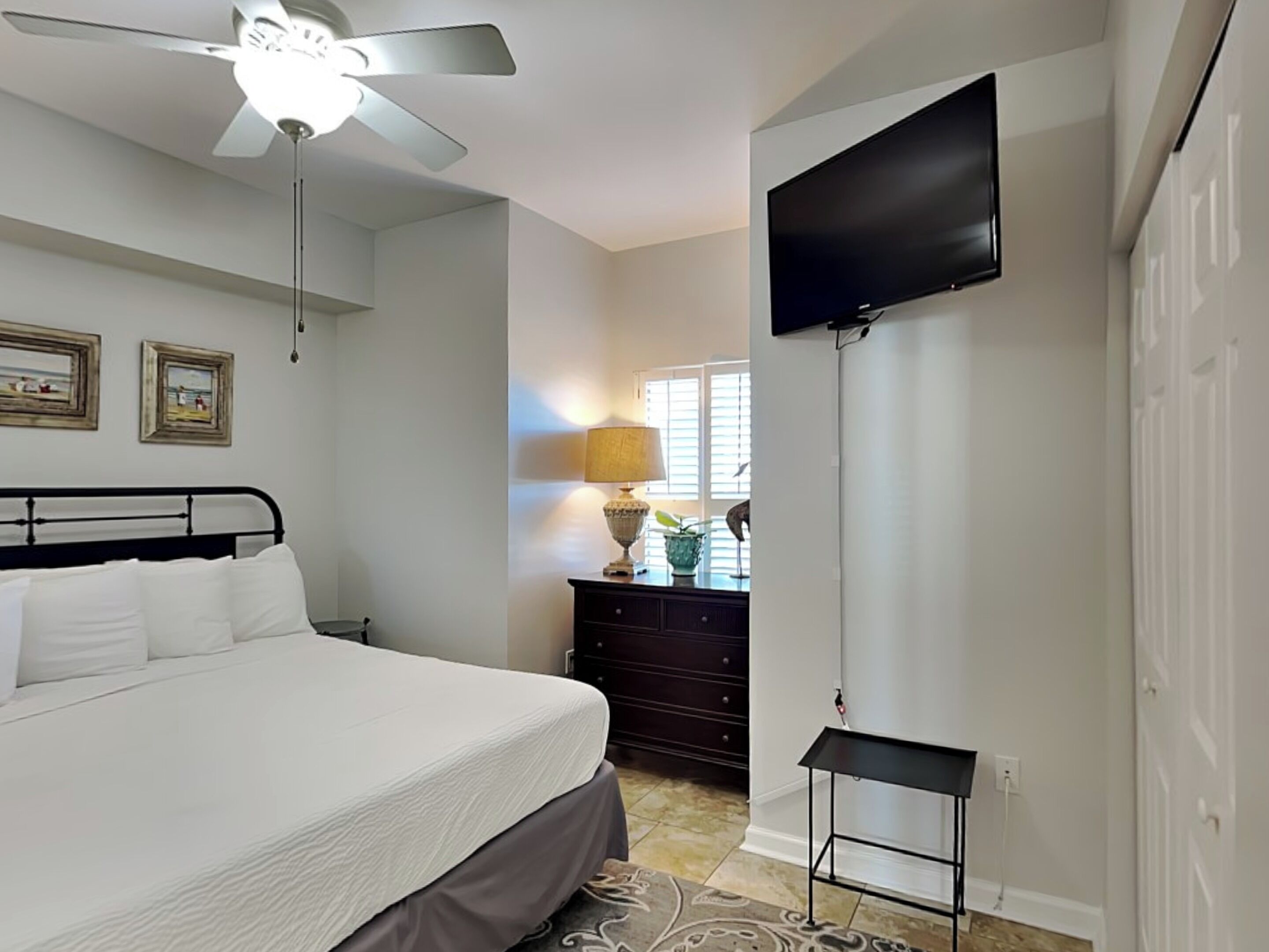 A bedroom with a bed, a ceiling fan, a TV mounted on the wall, a dresser, and a lamp.