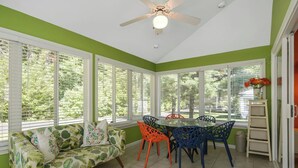 A sunroom with a ceiling fan, large windows, a dining table with colorful chairs, and a sofa with tropical prints. - Cottages at Summer Village, fully furnished cottage, Wi-fi, heated pools, hot tub (Wells)