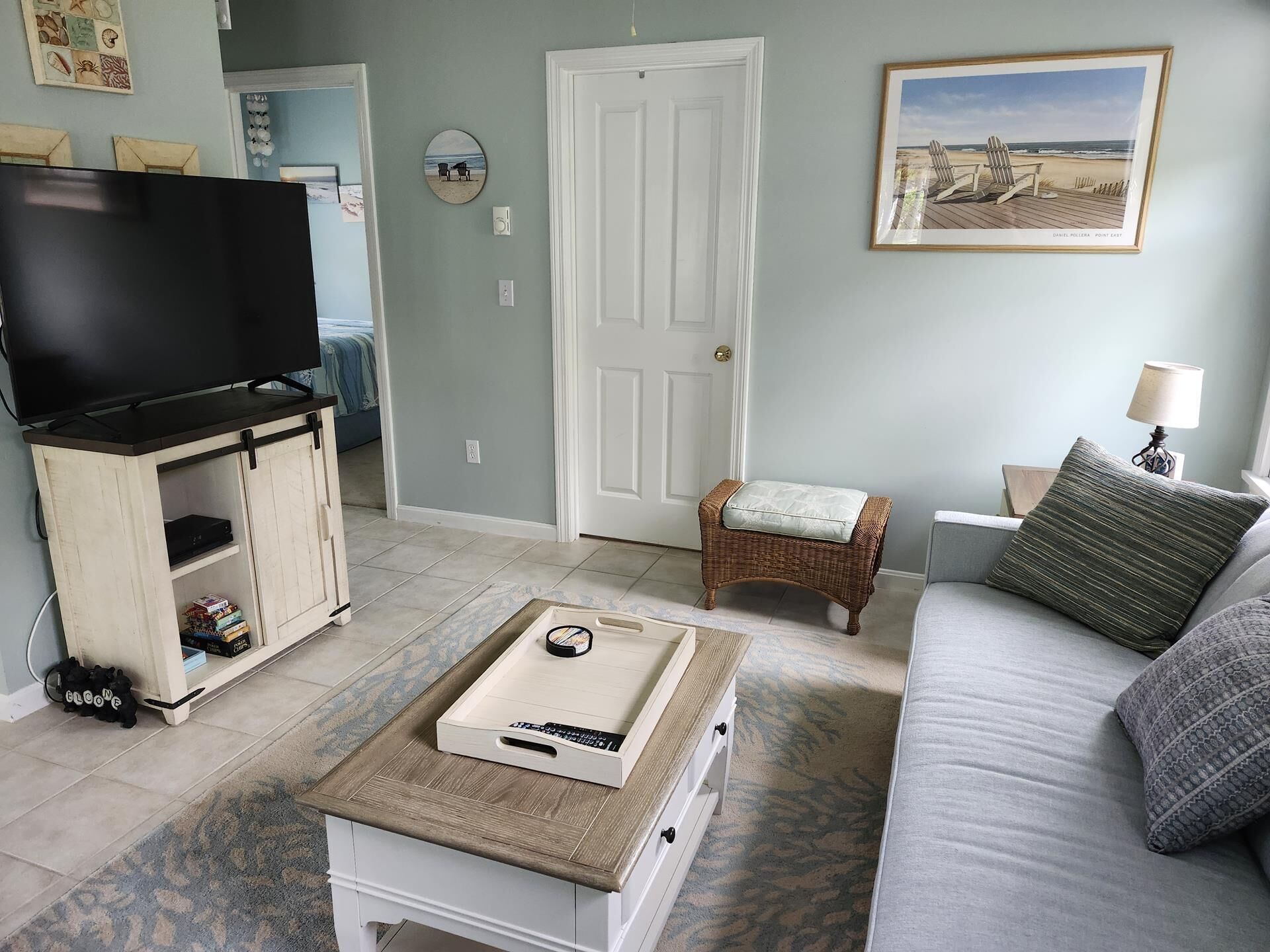 Una sala de estar con una televisión sobre un mueble bajo, una mesita con un control remoto, un sillón de mimbre, un sofá y una lámpara.