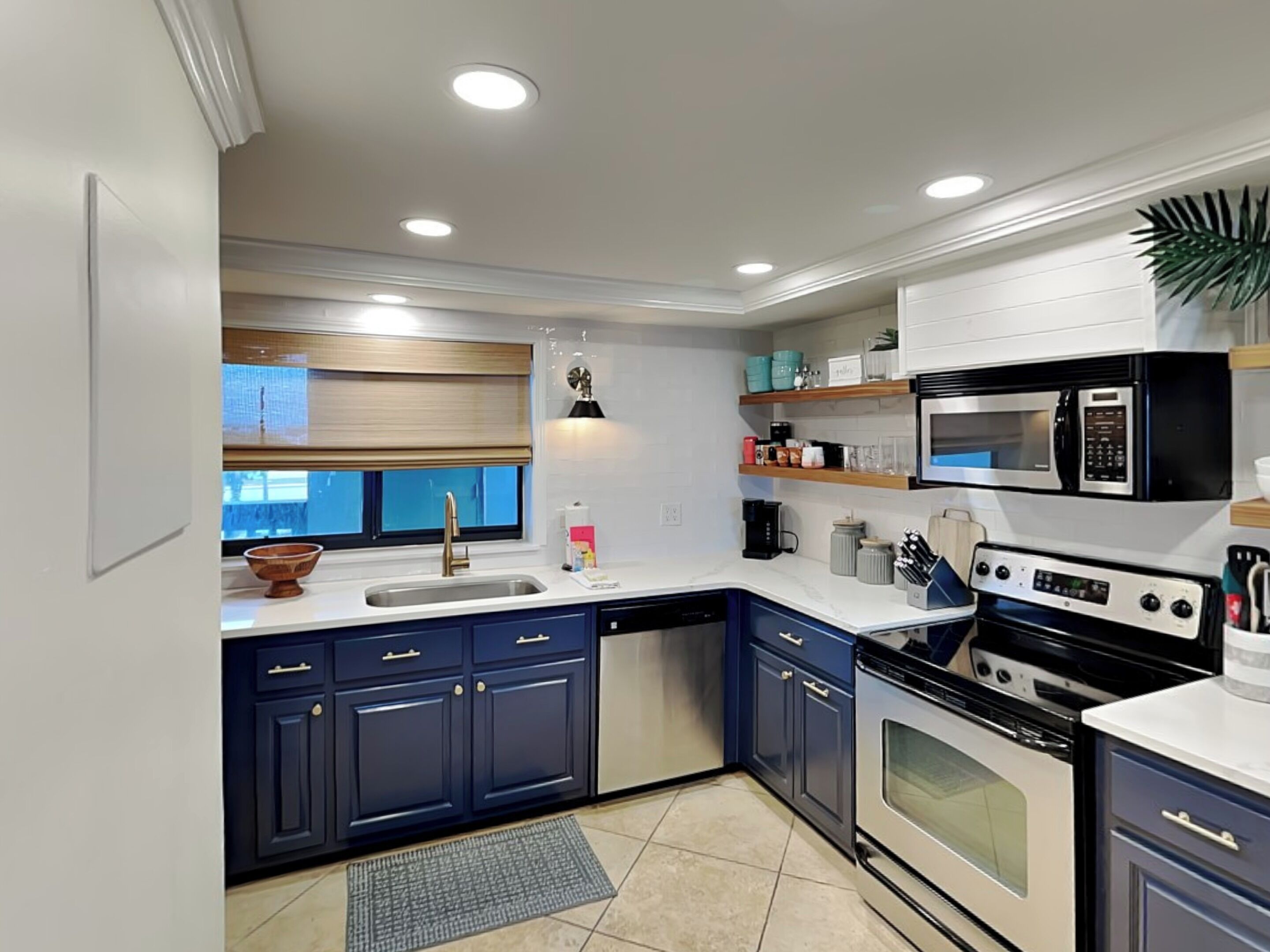 A modern kitchen with dark blue cabinets, a stainless steel sink, a microwave, a stove, and a window with blinds.