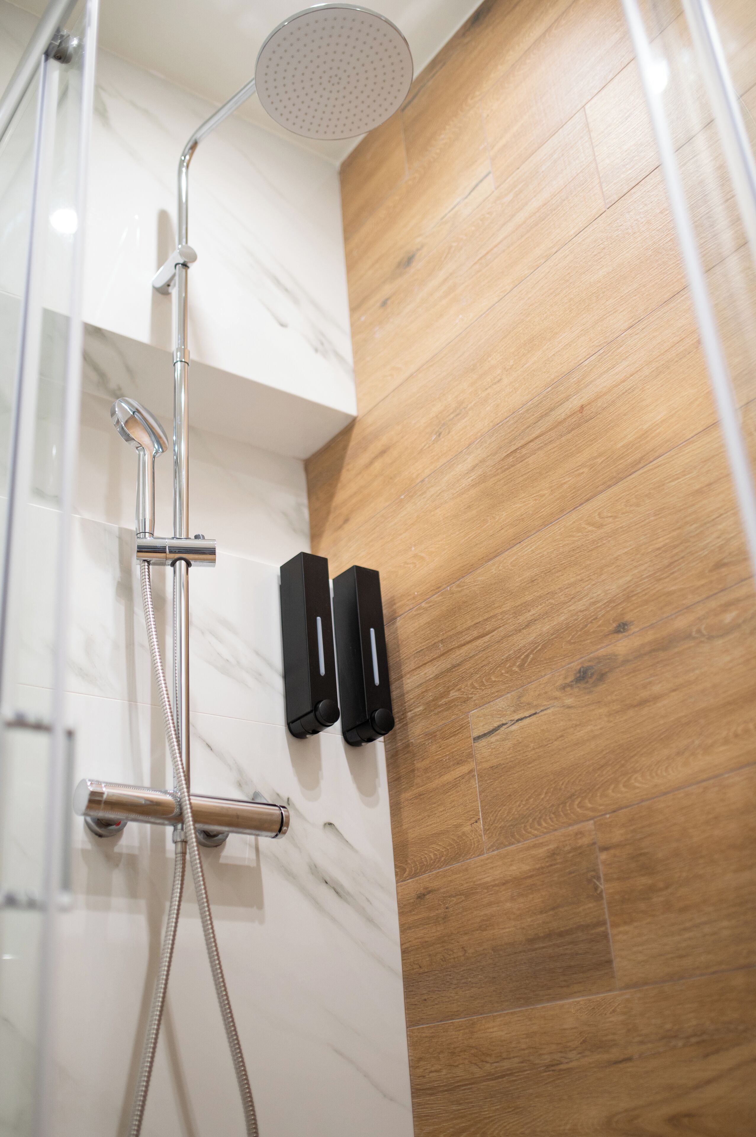 Ducha con cabezal de ducha manual y panel de control en la pared, suelo de madera y pared de mármol.