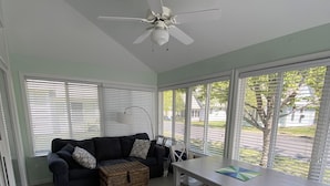 Una habitación con ventilador de techo, sofá oscuro con cojines estampados, una cesta de mimbre, una mesa blanca con posavasos verde y ventanas amplias con persianas.