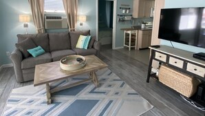 Una sala de estar con ventilador de techo, un sofá con cojines, una mesa de centro con una cesta, un televisor sobre un mueble bajo, y una cocina al fondo.