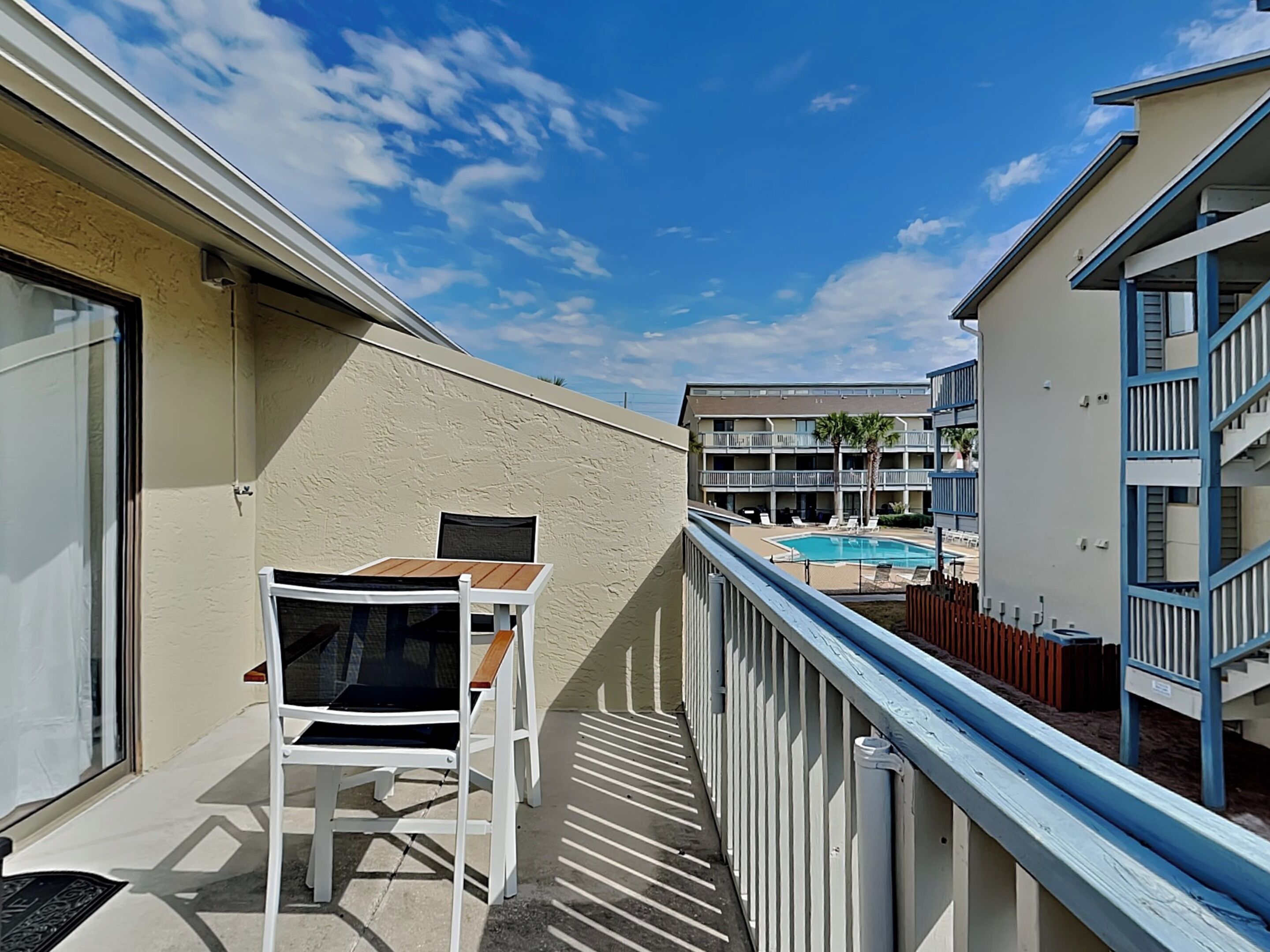 Un balcon avec une chaise blanche et une petite table, donnant sur une piscine et les bâtiments adjacents.