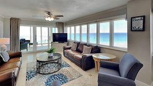 A spacious living room with a large window overlooking the ocean, a ceiling fan, a flat-screen TV, and a comfortable seating area. - Dunes of Panama D by Resort Collection (Panama City Beach)