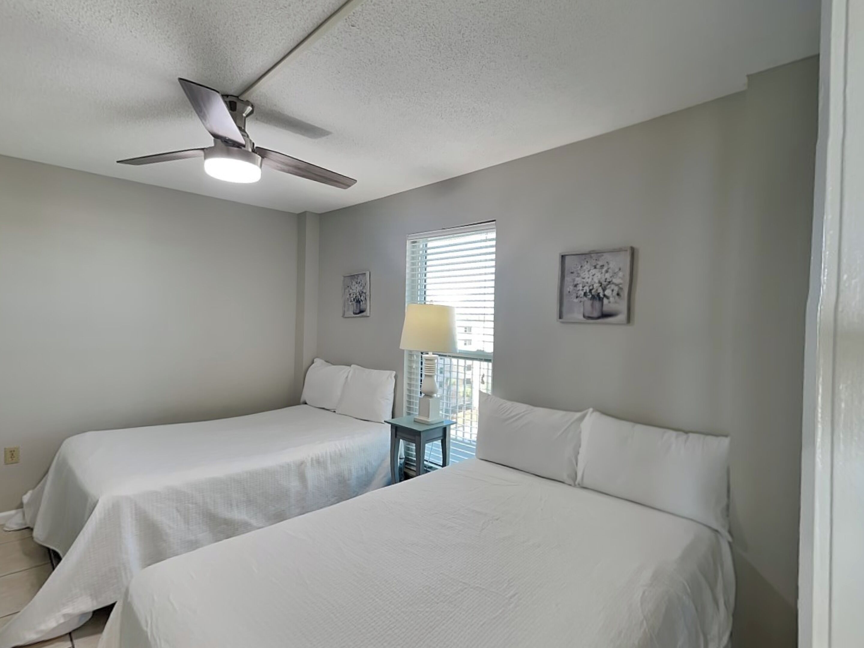 Une chambre d’hôtel avec deux lits simples, un ventilateur de plafond, une fenêtre avec des stores et une petite table avec une lampe.