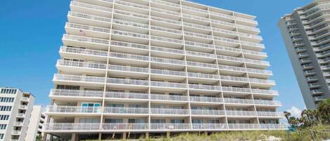 A multi-story hotel with balconies, located near a sandy beach.