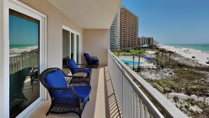 A balcony with two wicker chairs and a table, overlooking a beach and a hotel building. - Dunes of Panama D by Resort Collection (Panama City Beach)