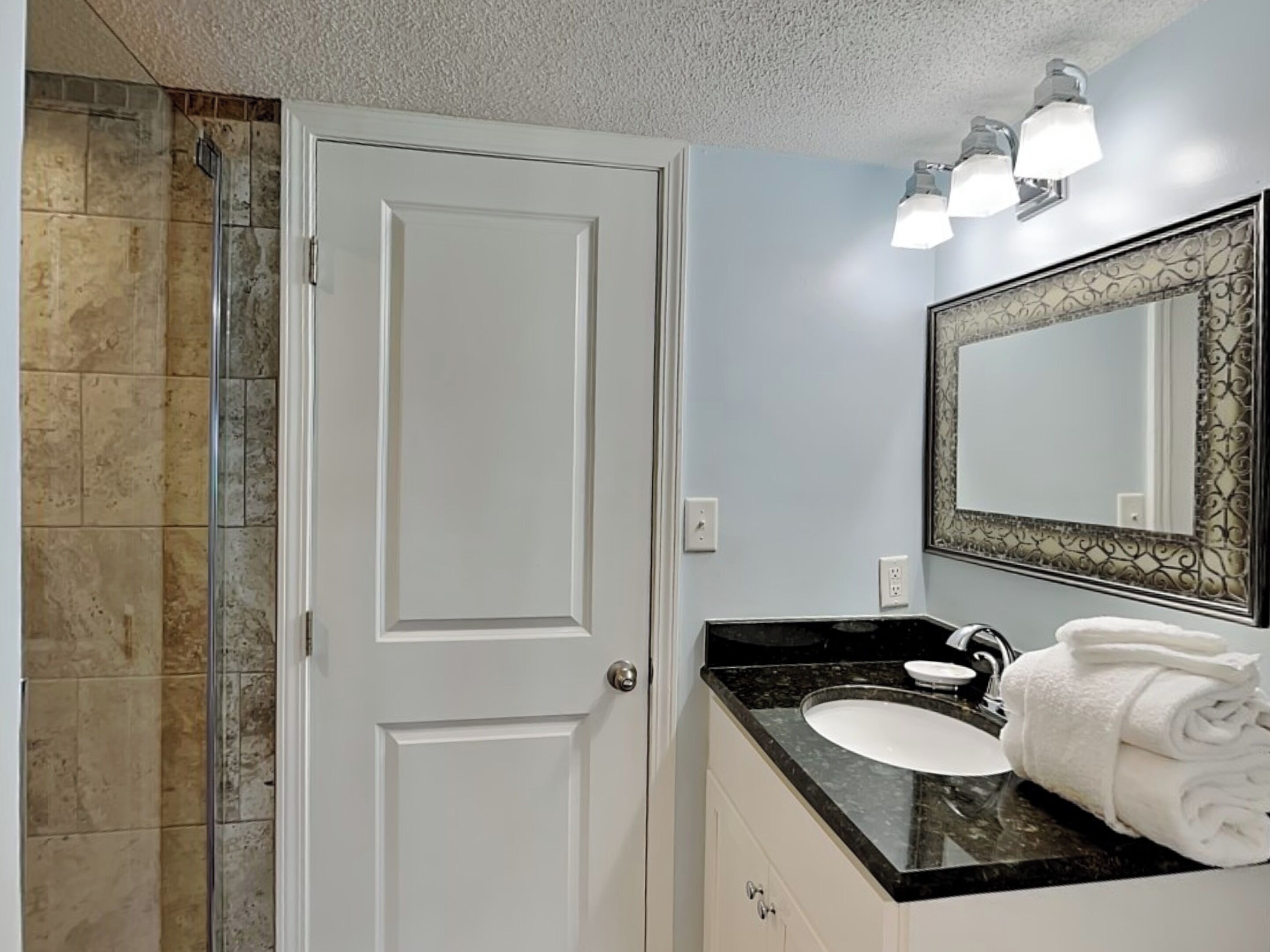 Un baño con una puerta blanca, un lavabo con encimera negra y un espejo con marco decorativo.