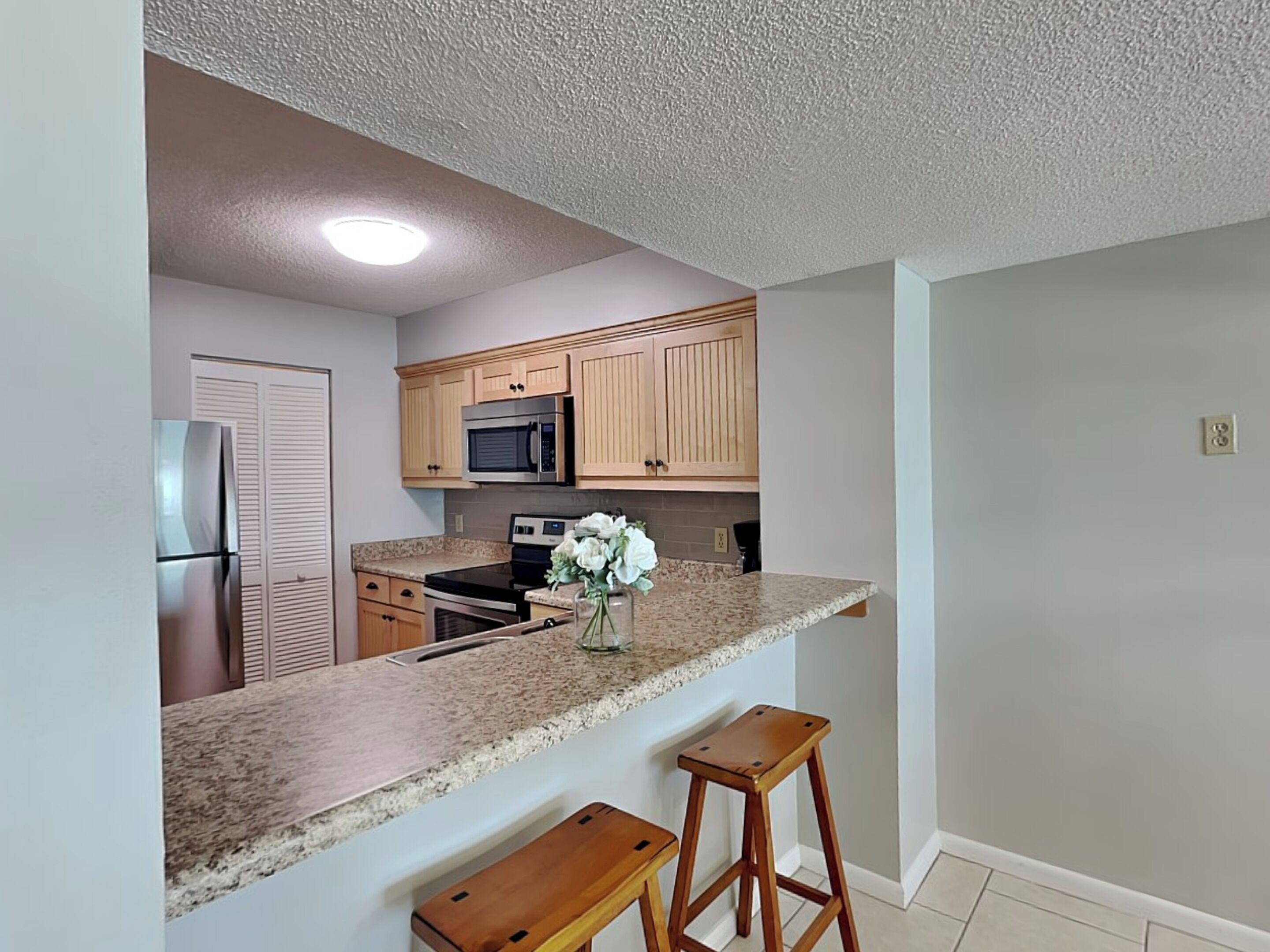 A kitchen with wooden cabinets, a stainless steel refrigerator, a microwave, and a granite countertop. There are two wooden stools and a vase with flowers on the counter.