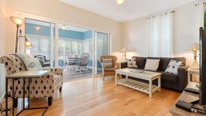 A living room with a sofa, chairs, a coffee table, and a TV. There is a sliding glass door leading to a balcony. - Cottages at Summer Village, fully furnished cottage, Wi-fi, heated pools, hot tub (Wells)