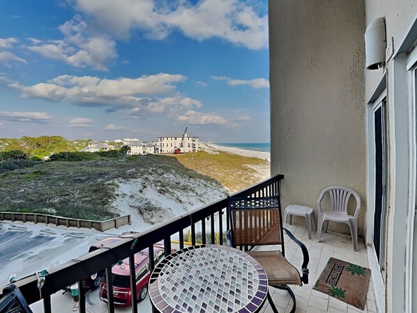 A balcony with a view of the beach and a building in the distance. There is a round table with a chair and two plastic chairs.