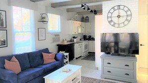 Un salon moderne avec un canapé bleu, une table basse blanche et un téléviseur posé sur un meuble blanc. On y trouve également un coin cuisine avec des meubles blancs et une horloge au mur.