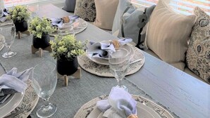 Un coin repas avec une table dressée pour quatre personnes, agrémenté d’un lustre, de grandes fenêtres équipées de stores et offrant une vue sur un bâtiment en briques.