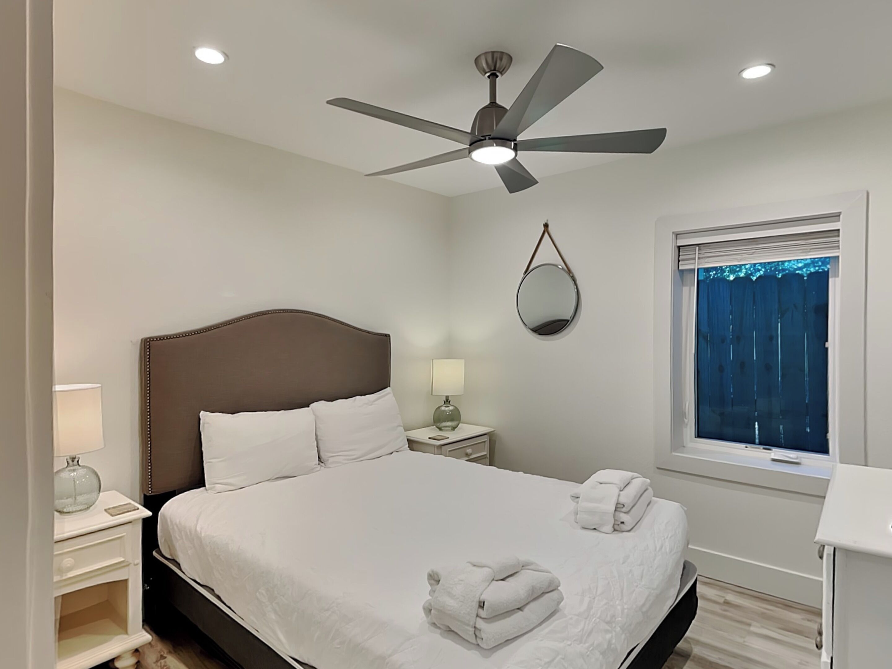 A neatly arranged bedroom with a large bed, a ceiling fan, a bedside table with a lamp, a mirror, and a window with blinds.