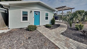 Una casa de un piso con puerta azul, un porche cubierto y un jardín arbolado con arbustos pequeños y una palmera.