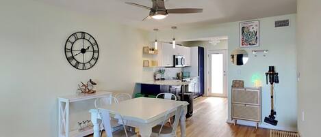 Un comedor amplio con una mesa y sillas blancas, una cocina al fondo y un ventilador de techo.