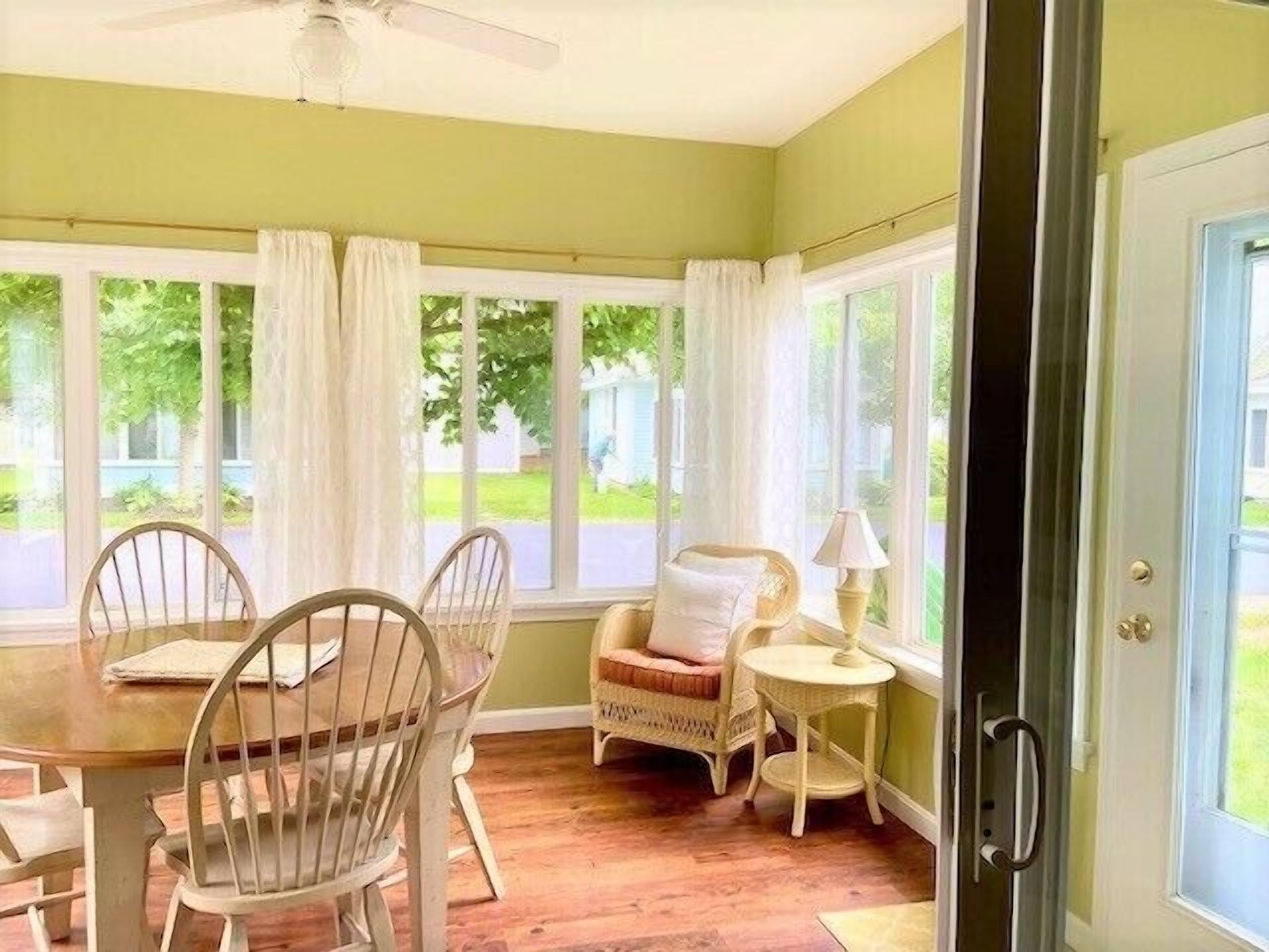 A sunroom with a dining table, chairs, a wicker chair, a small table with a lamp, and large windows with curtains.