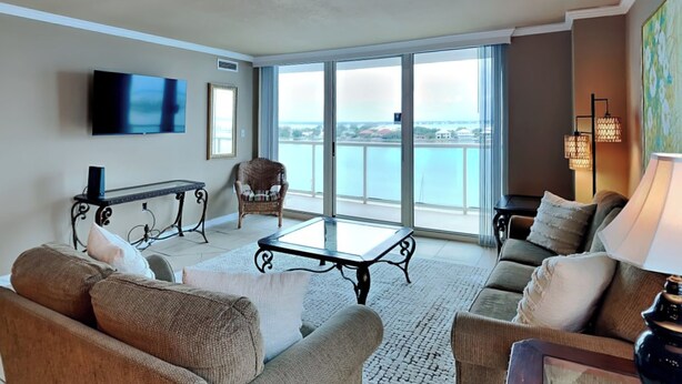 Un vaste salon doté d’une grande fenêtre offrant une vue sur la mer, d’un téléviseur à écran plat fixé au mur et d’un coin salon confortable avec des canapés et une table basse.