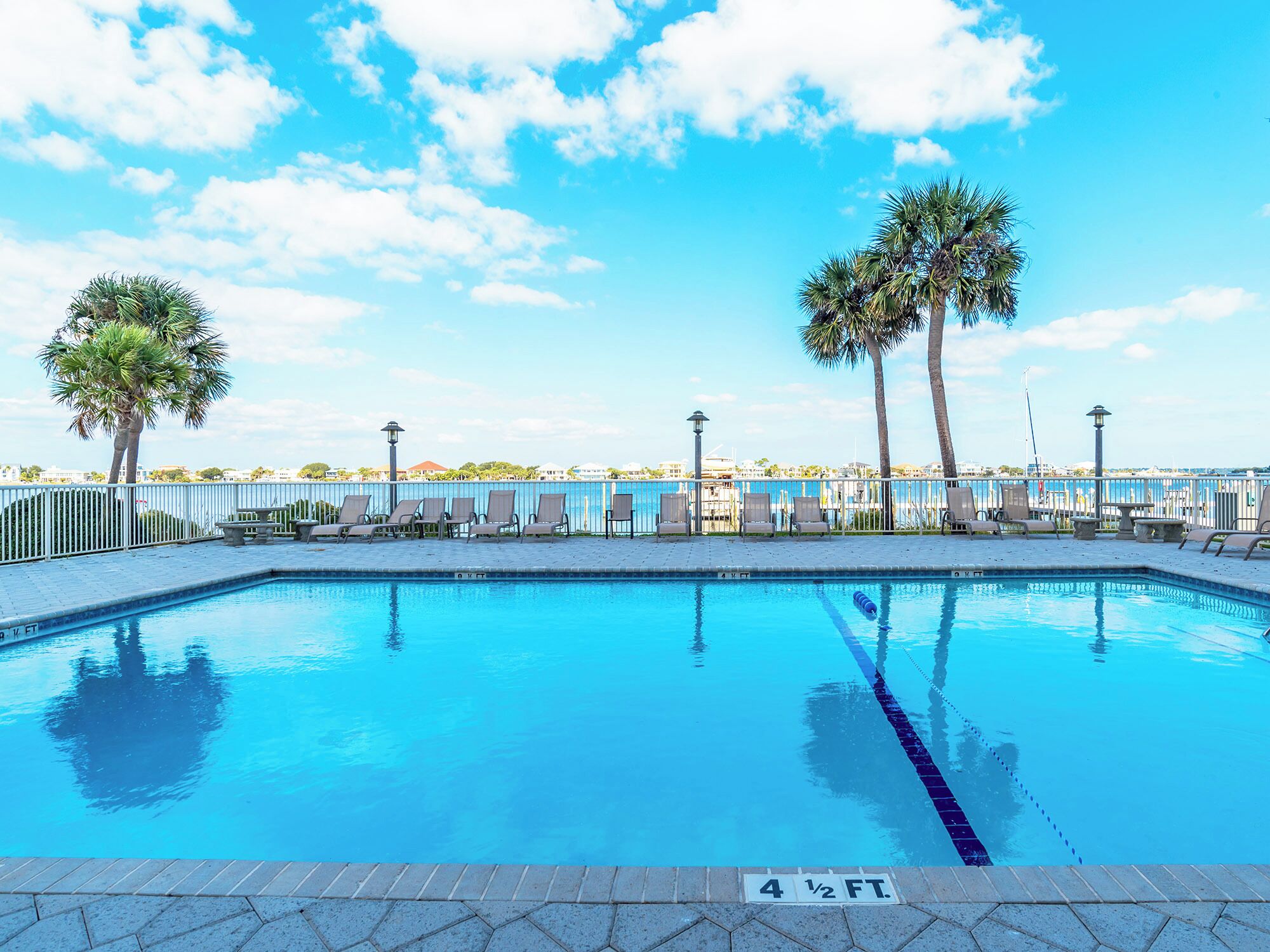 A rectangular swimming pool with a 4 1/2 ft depth, surrounded by lounge chairs and palm trees, with a clear view of the water and a distant shoreline.