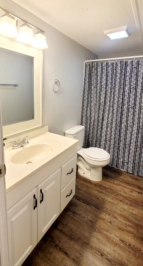 Une salle de bain avec un lavabo, un meuble-lavabo et des toilettes, tous de couleur blanche. Il y a un rideau de douche avec un motif noir et blanc.
