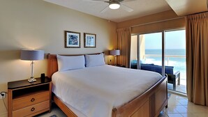 Ein Hotelzimmer mit einem großen Bett, zwei Nachttischlampen, einem Nachttisch, einem Holzschrank, einem blauen Teppich und Meerblick durch eine Schiebetür.