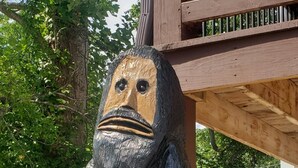 Eine hölzerne Skulptur einer Gestalt mit großem Kopf und langem Körper, draußen vor einer Holzkonstruktion inmitten von Grünpflanzen.