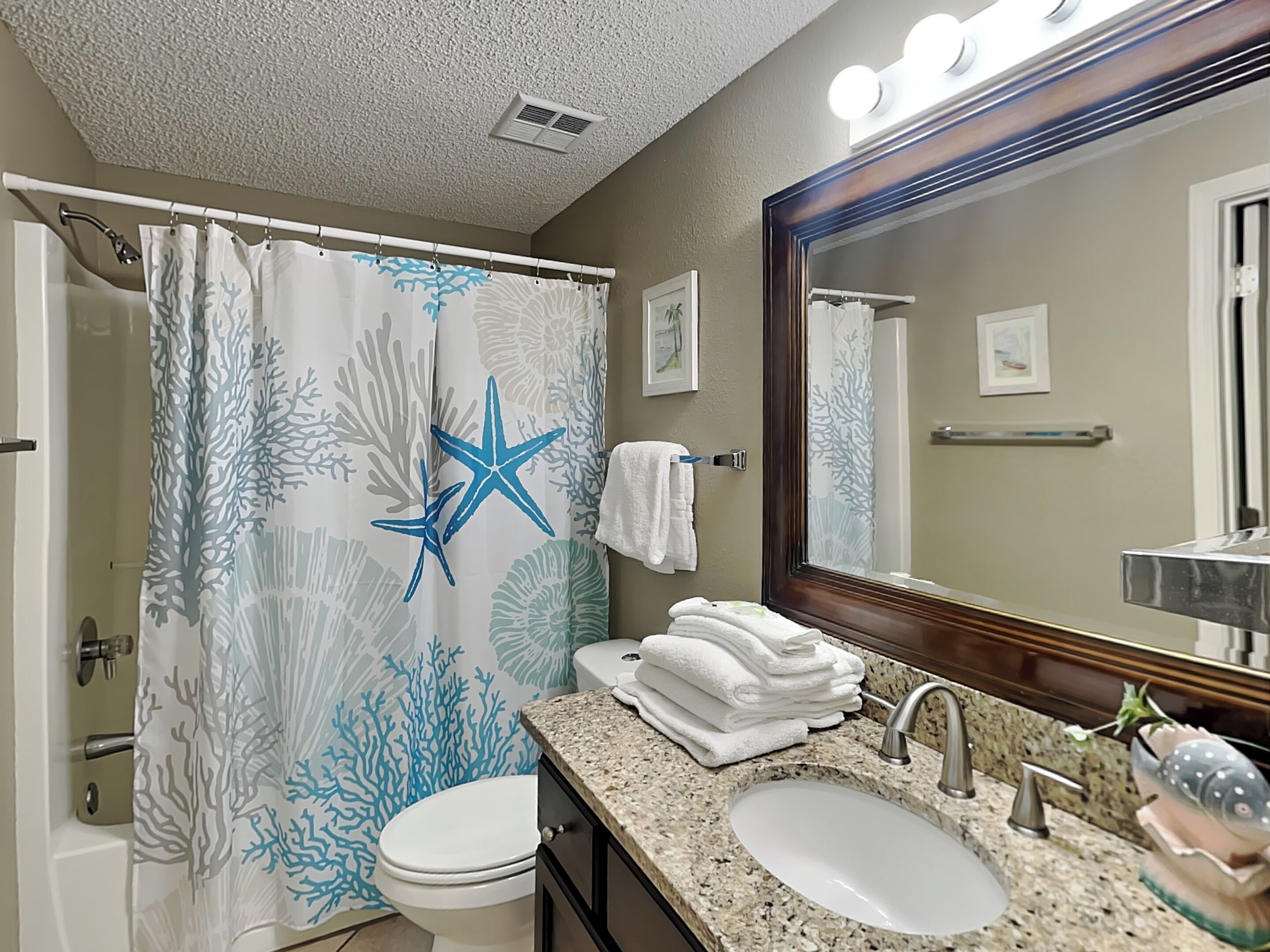 Une salle de bain avec un rideau de douche orné d’une étoile bleue et de motifs coralliens, des toilettes blanches, un lavabo avec un miroir au-dessus, et un porte-serviettes avec des serviettes pliées.