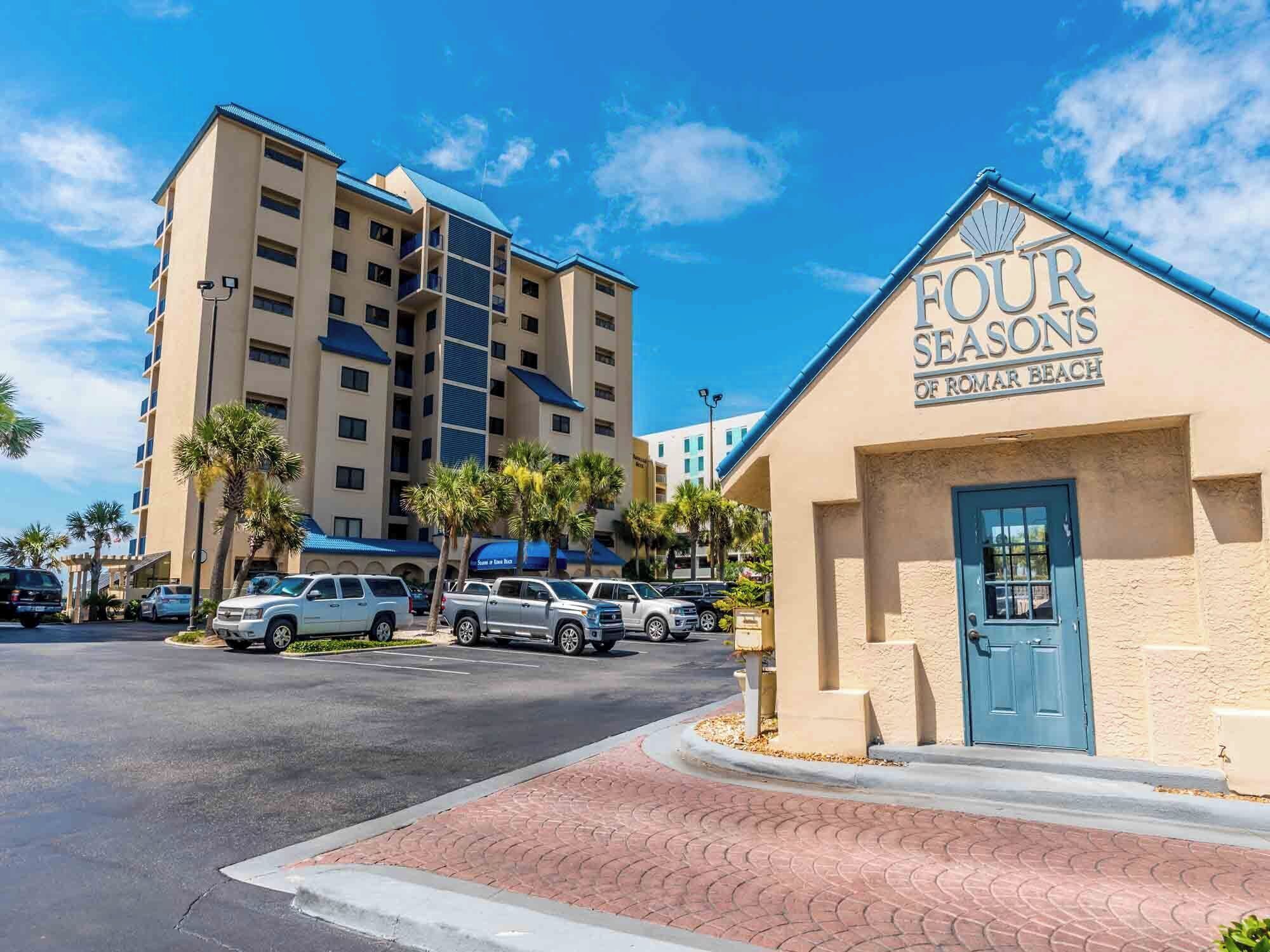Un hotel llamado 'Four Seasons of Romar Beach' con una puerta azul, coches aparcados y un aparcamiento.