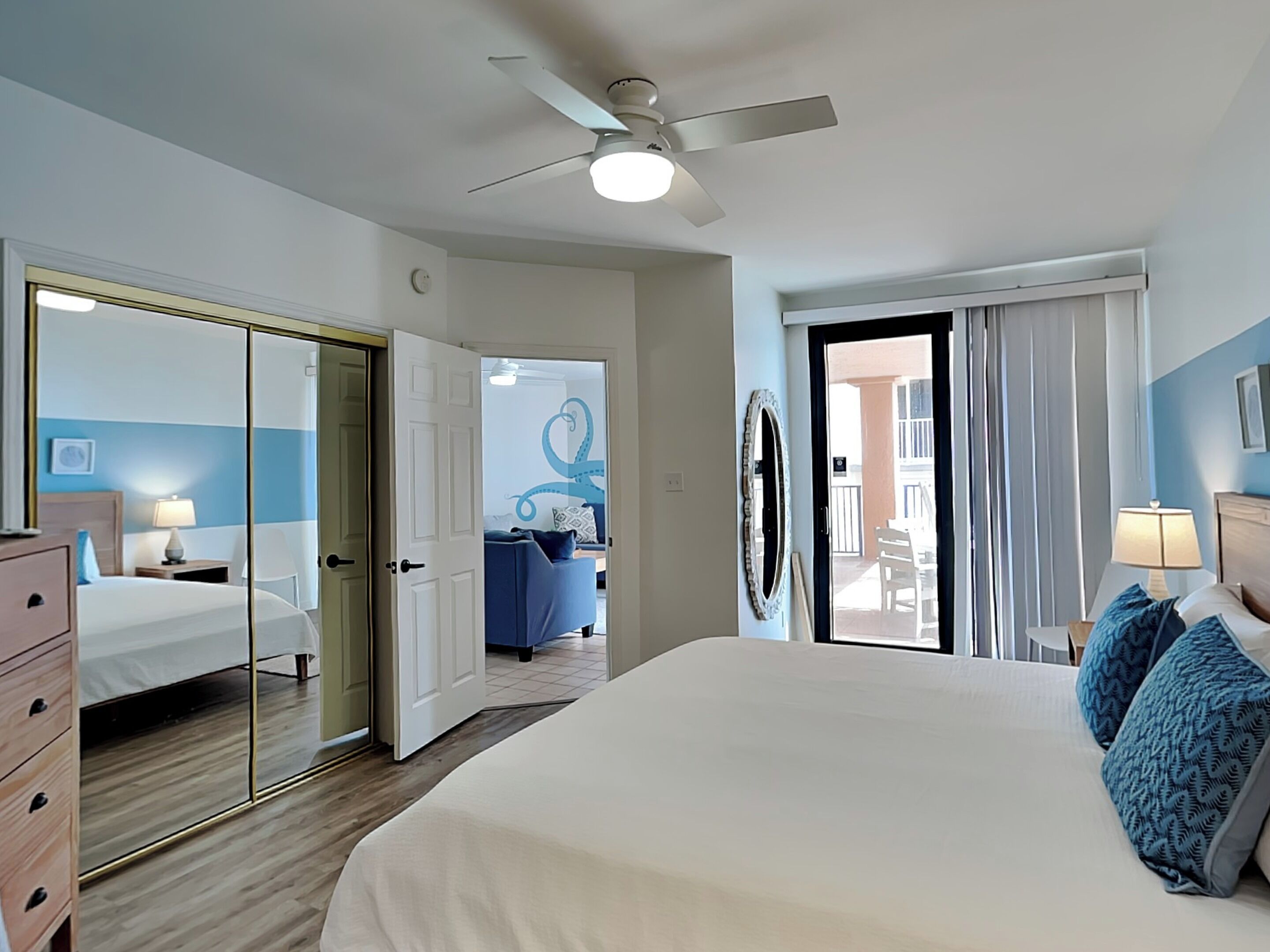 Une chambre d’hôtel avec un grand lit, un ventilateur de plafond, une porte donnant sur un balcon et une vue sur l’extérieur.