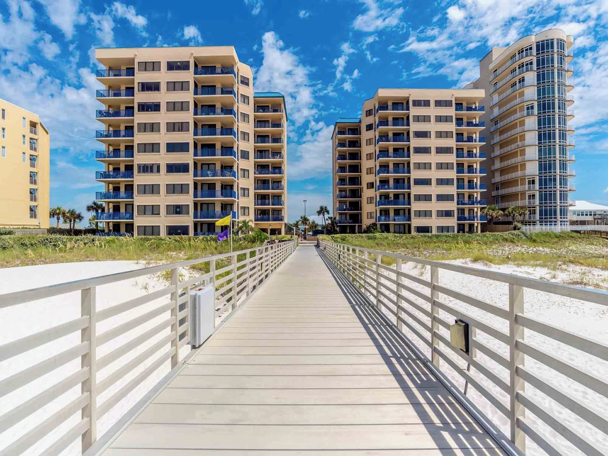 A boardwalk leading to beachfront buildings with balconies, a clear sky, and a sandy beach.