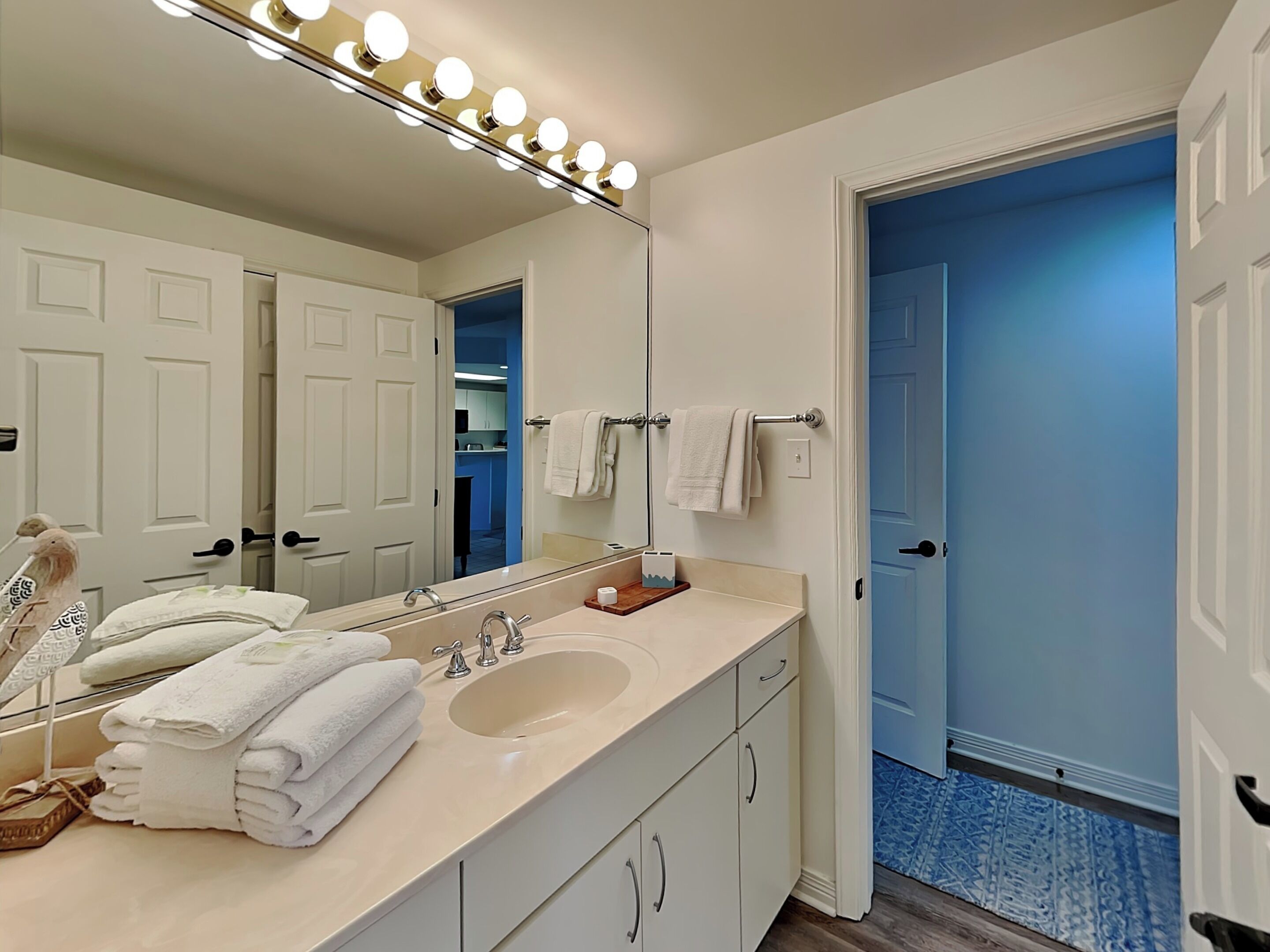 A bathroom with a vanity, sink, and mirror with lights. There are towels stacked on the vanity and a door leading to another room.