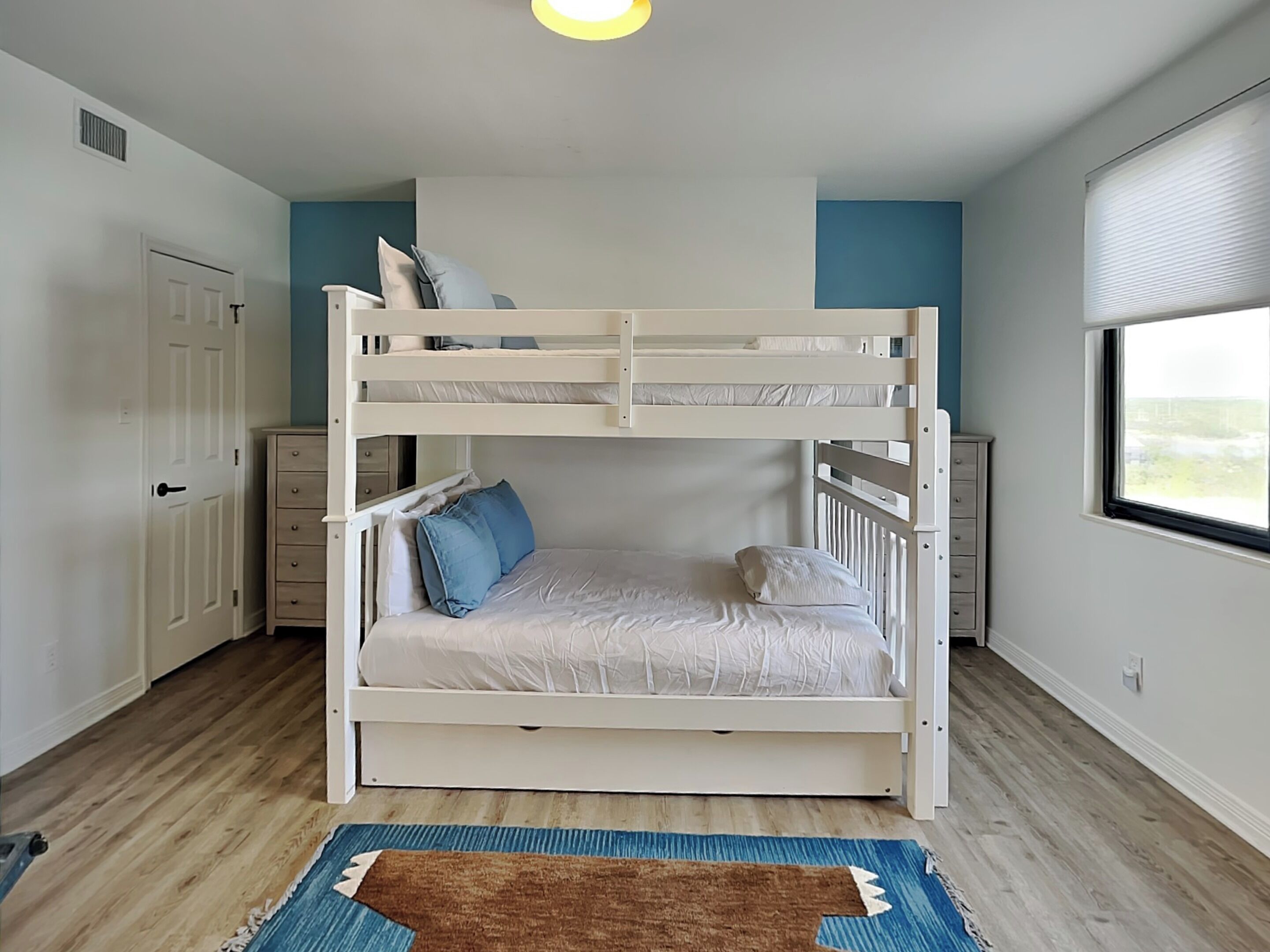 A bunk bed with white bedding and blue pillows, a wooden floor, a blue and brown rug, a dresser, and a window with a view.