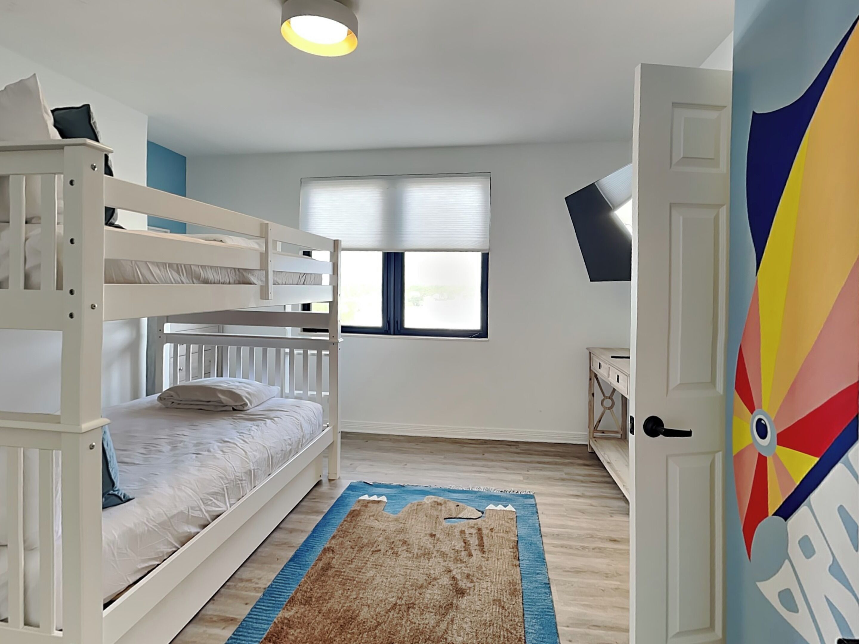 Una habitación con literas, una ventana con persianas, una mesita pequeña y una puerta colorida.