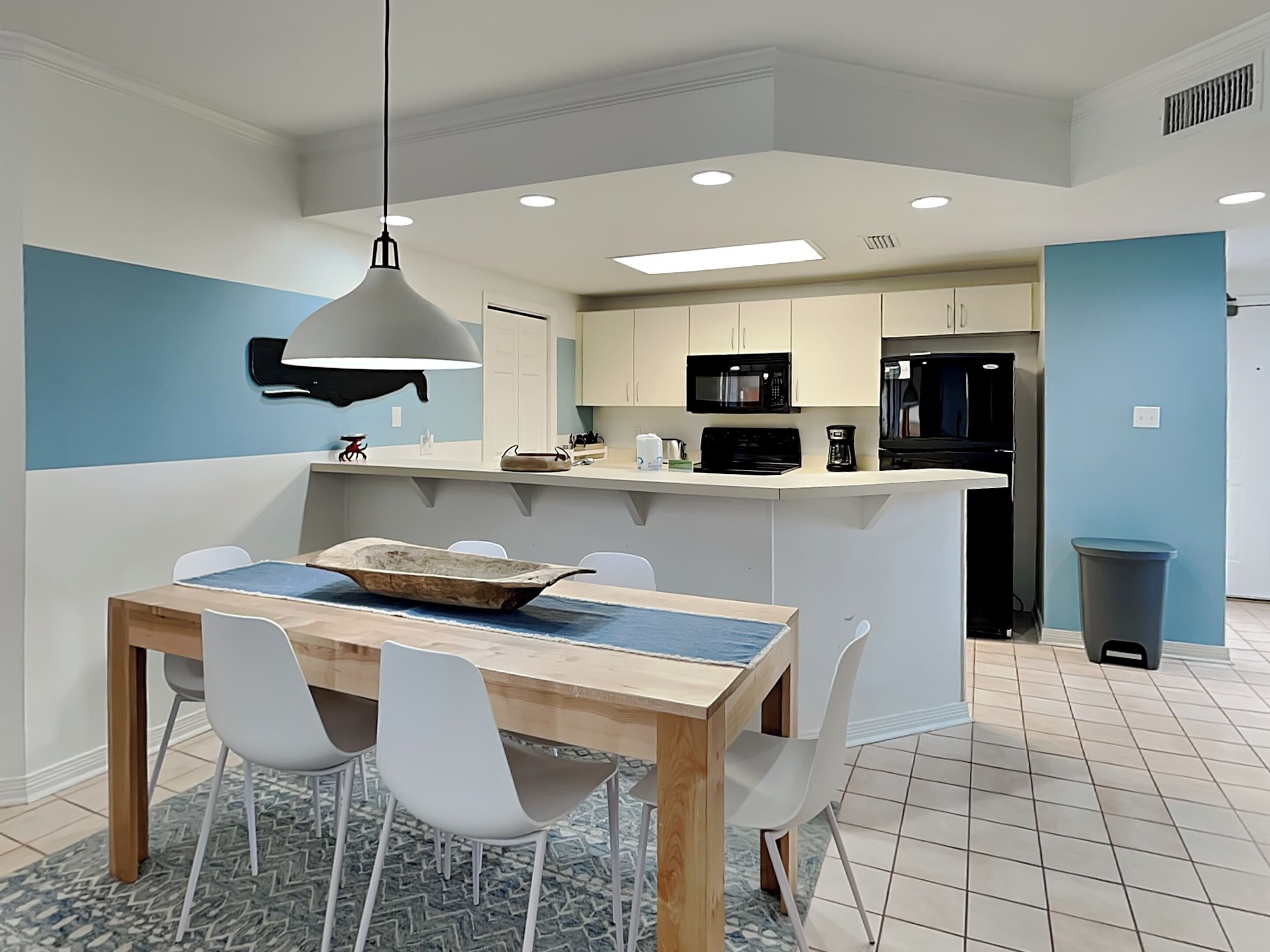 A modern kitchen with white cabinets, a black microwave, and a black refrigerator. There is a dining area with a wooden table and white chairs.