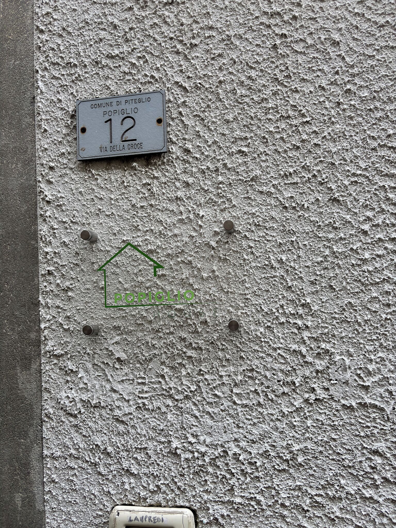 A textured wall with a sign indicating 'COMUNE DI PITEGLIO POPIGLIO 12 VIA DELLA CROCE' and a green house symbol.