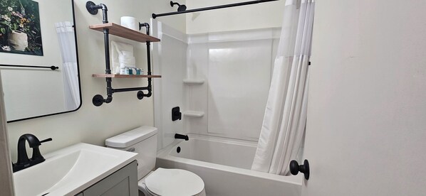 Un baño moderno con lavamanos, inodoro y bañera blancos. Hay un estante con artículos de aseo y un espejo sobre el lavamanos.