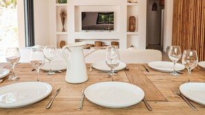 A dining area with a wooden table set for a meal, white plates, and wine glasses. A TV is mounted on the wall in the background.