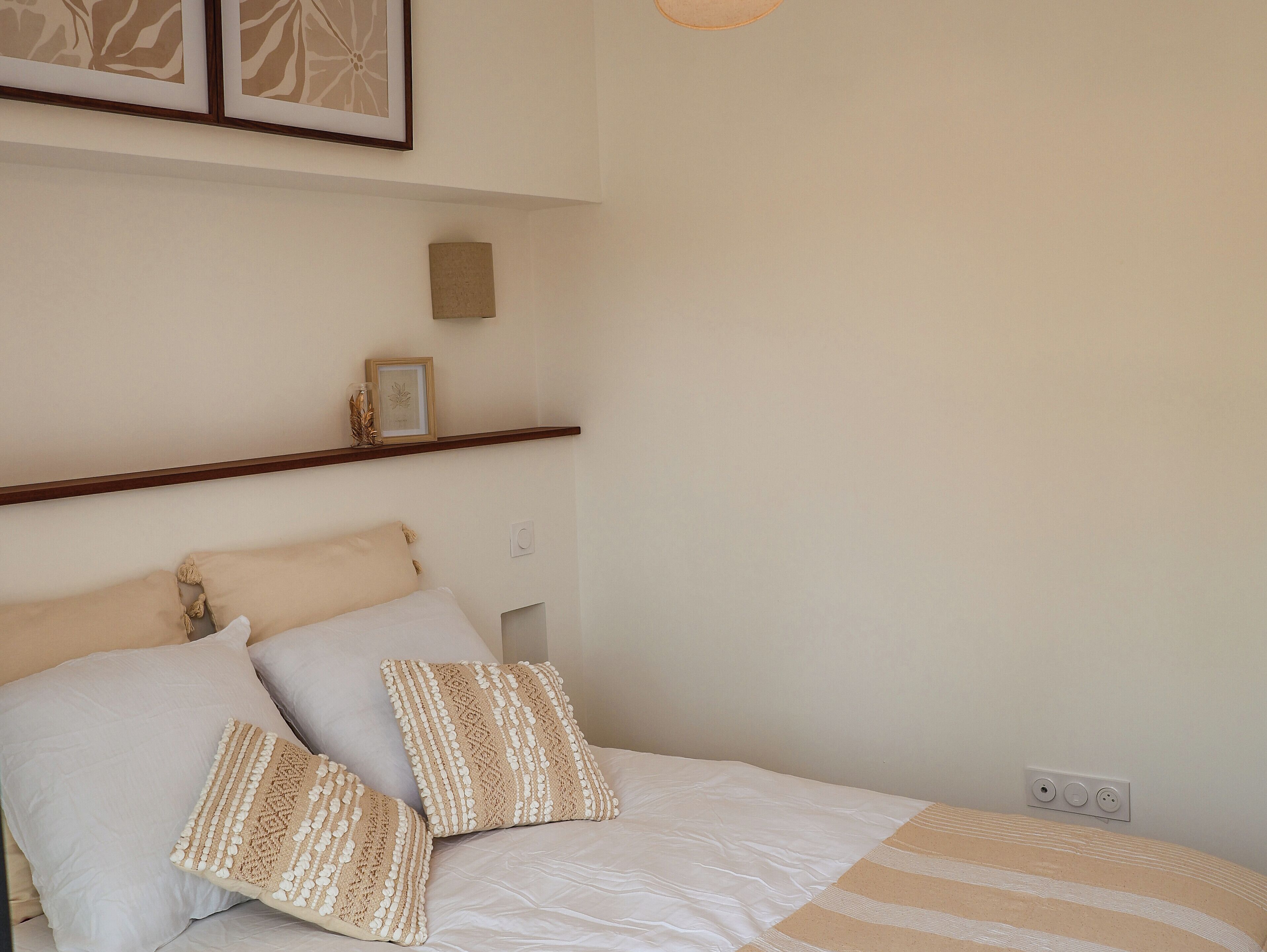 A neatly made bed with a striped blanket and two decorative pillows, a wooden shelf with a framed picture and a small decorative item, and a power outlet on the wall.