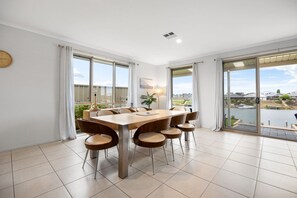 Een eethoek met een grote tafel en stoelen, grote ramen met gordijnen, en uitzicht op een waterpartij en gebouwen.
