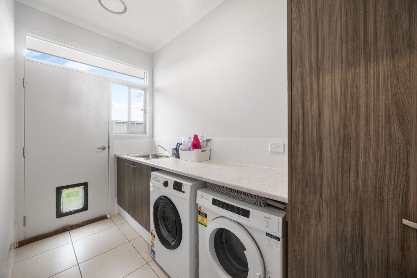 A compact laundry room with two front-loading washing machines, a countertop with a sink, and a window providing natural light.