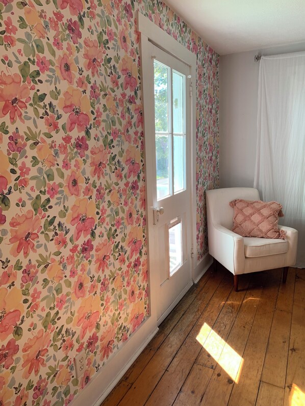 Una habitación con papel tapiz floral, una puerta blanca, un sillón blanco con cojín y suelo de madera.