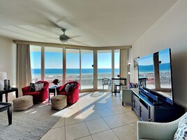A spacious balcony with a view of the ocean, furnished with red chairs and a table, and a ceiling fan.